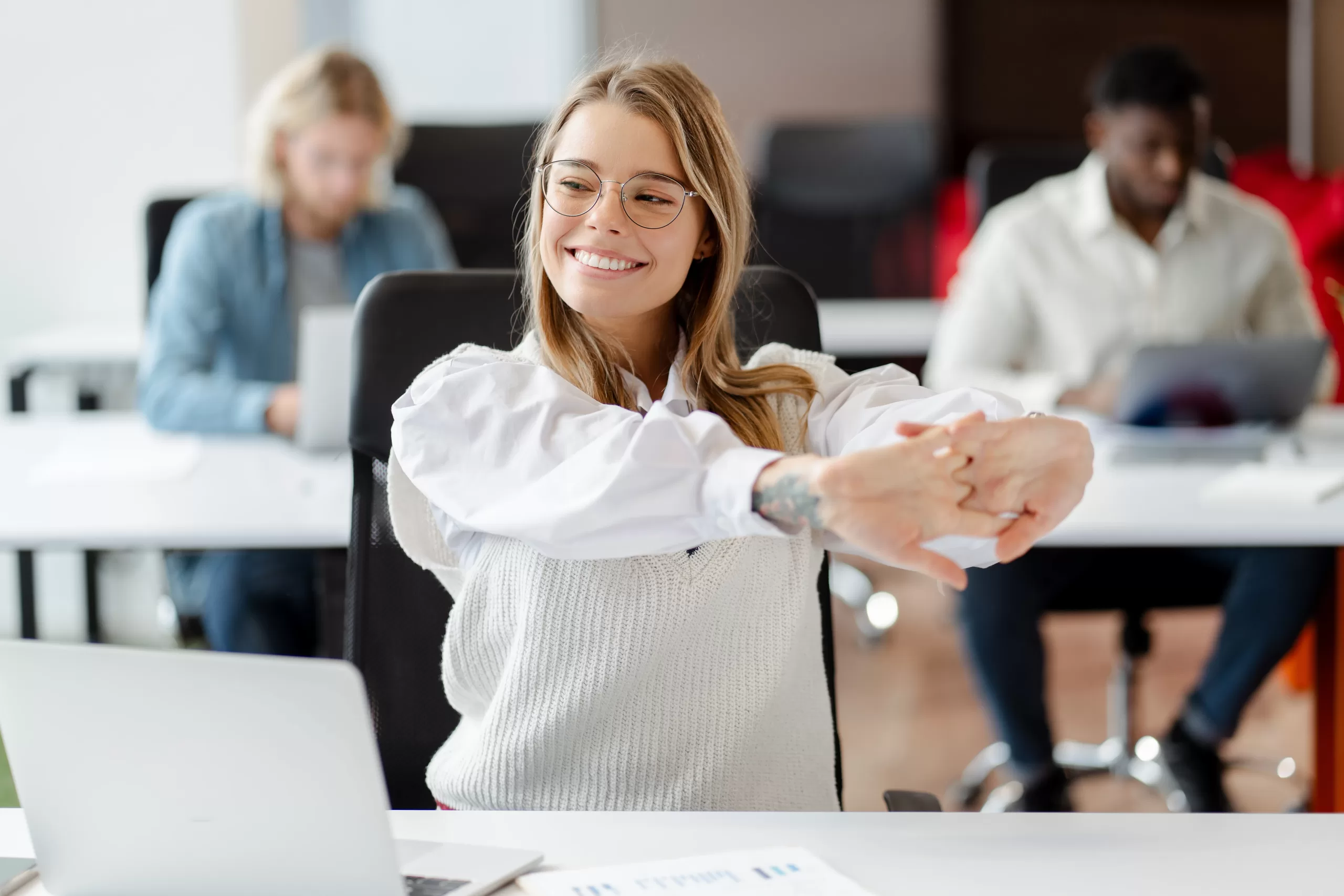 Perfect posture - A woman sitting in an office environment, stretching her arms out in front of her with linked fingers. Smiling.