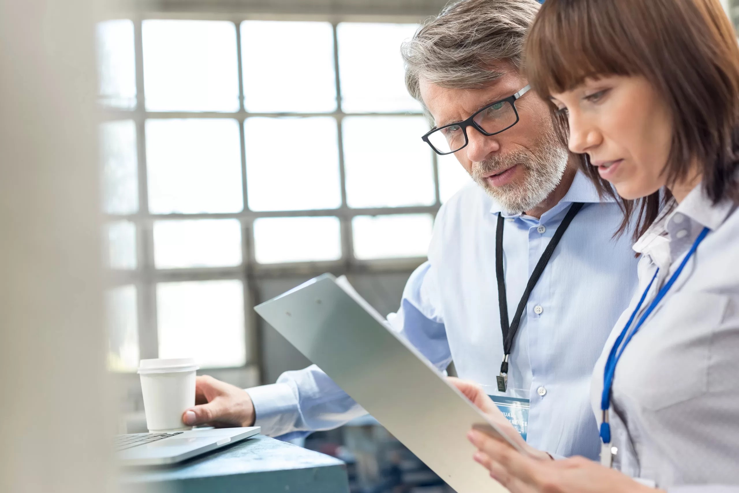 Occupational Health Support - Two work colleagues studying a clipboard. Internal office environment, daylight through windows in background.